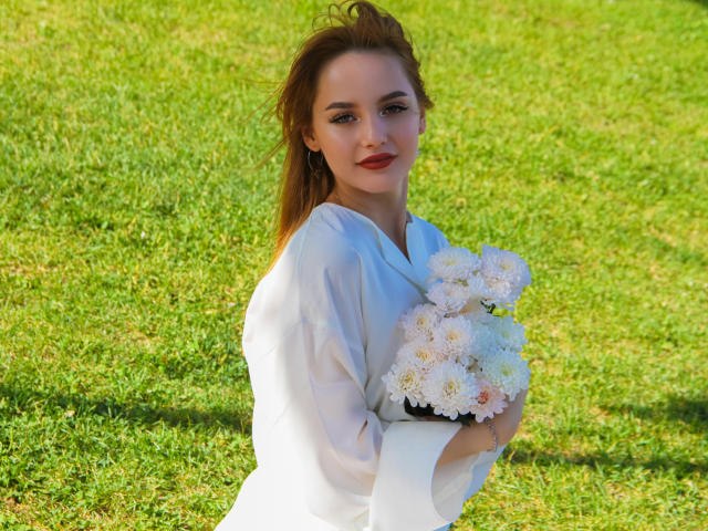 CamillaRoberts with white dress, holding white chrysanthemums, has red lipstick, and is standing on green grass, with brown hair in a ponytail. Her tits are small and cute.