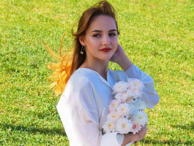 CamillaRoberts with orange hair, white blouse, holding bouquet of white and pink flowers, grassy background, hand in hair, red lipstick, earrings, smiling, sunlight.