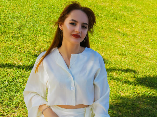 CamillaRoberts with red hair, white blouse, and white pants, sitting on green grass, smiling, wearing red lipstick, small hoop earrings, and a bracelet.