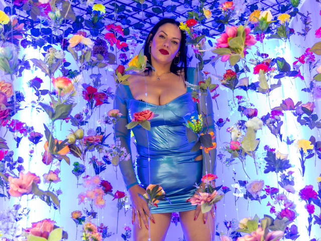 LizaRussell with big tits in a shiny blue dress, standing in front of a colorful flower wall, smiling with red lipstick, dark hair, and a cute face.