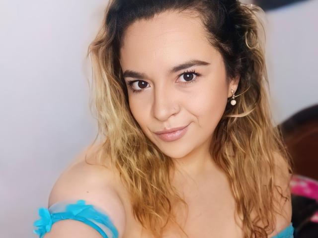 NaugthyCurvy hat welliges blondes Haar, braune Augen und ein süßes Lächeln. Sie trägt ein blaues Oberteil mit einem Schulterauschnitt, das ihre Brüste zeigt, und trägt eine Perlenohrring in ihrem rechten Ohr..