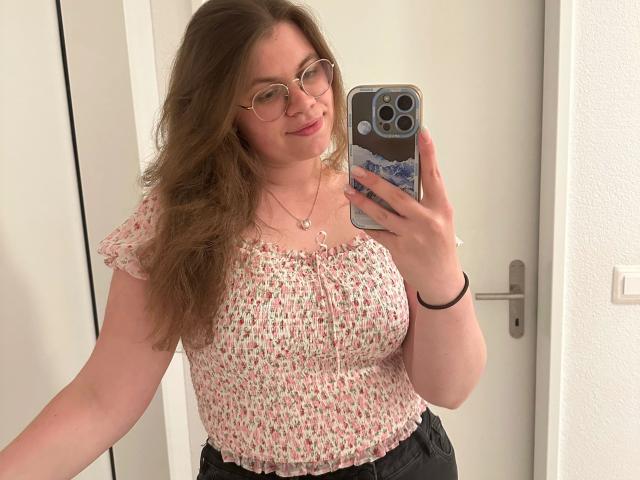 LinaLove-hot with wavy brown hair, glasses, floral top, small tits, holding phone, mirror selfie, white door, black pants, necklace, bracelet, smiling, standing in bright room.