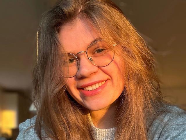LinaLove, com cabelos castanho-claros, óculos e um sorriso, vestindo um suéter cinza, banhada pela luz quente do sol. Seu traseiro não está visível, apenas seu rosto e parte superior do corpo..