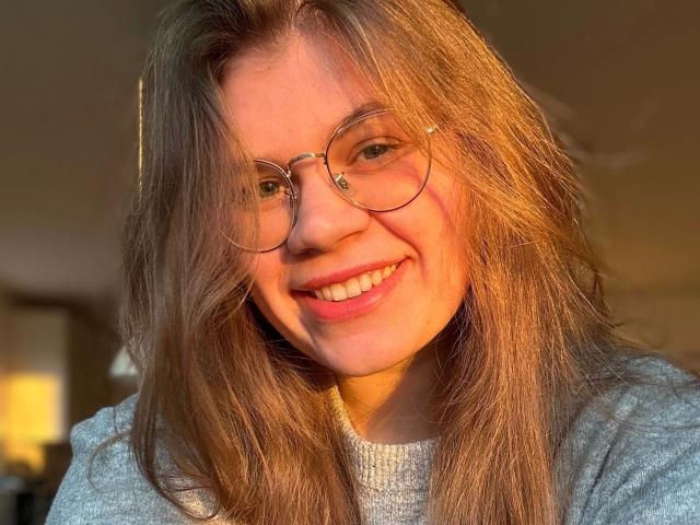 LinaLove, belle avec de longs cheveux bruns, souriante, portant des lunettes rondes et un pull gris, la lumière du soleil illuminant son visage, révélant son expression adorable et joyeuse, et ses lèvres douces de couleur rose pâle..