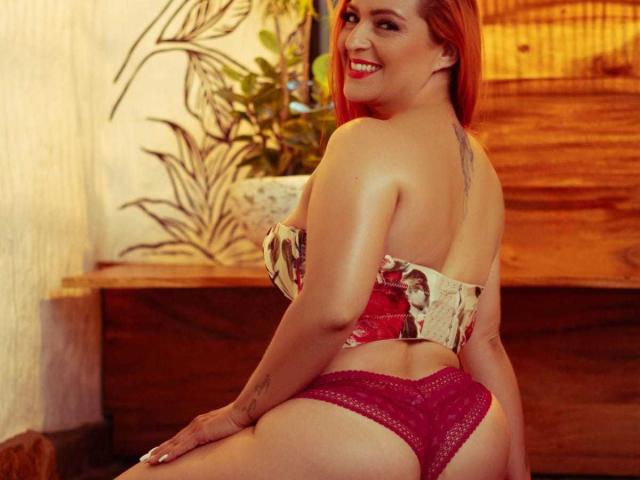Zoeray with red hair, smiling, wearing a floral strapless top and purple lace panties, showing her ass and a tattoo on her back. Her tits are partially visible.