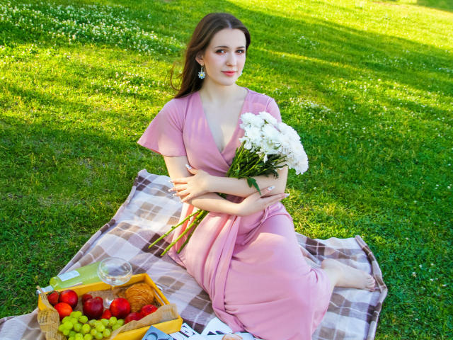 JuliaKing with tits covered by pink dress, sitting on checkered blanket, holding white flowers, green grass, basket with red apples and grapes, sunny day, earrings, bare feet.