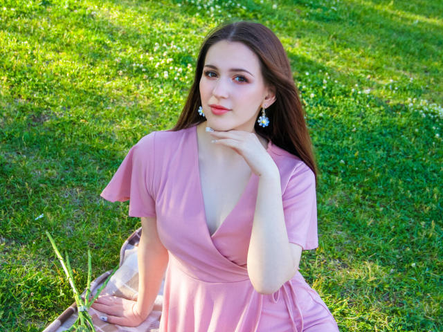 JuliaKing with brown hair, pink dress, and white earrings, sitting on grass, hand on chin, looking at camera, showing her tits, soft green background, sunny day.