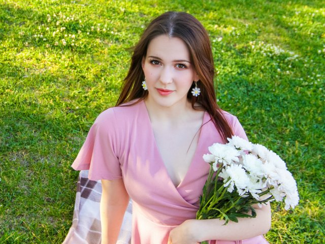 JuliaKing with long brown hair, pink dress, daisy earrings, holding white flowers, sitting on grass, looking up with a slight smile, sunny day, green grass.
