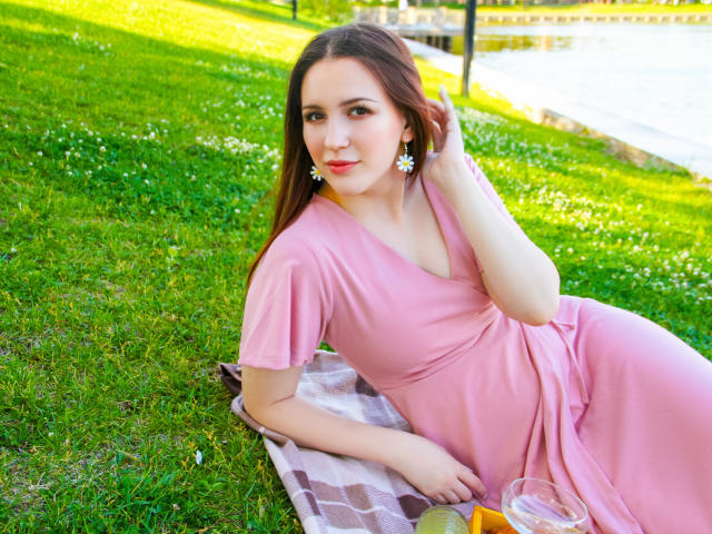 JuliaKing with long brown hair, pink dress, and earrings, lying on a checkered blanket, holding her hair, looking at the camera, with a grassy lawn and water in the background.