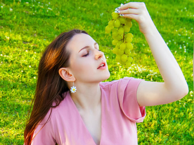 JuliaKing with brown hair, pink shirt, and daisy earrings, smelling green grapes held above her head. She's got her eyes closed and a slightly open mouth.