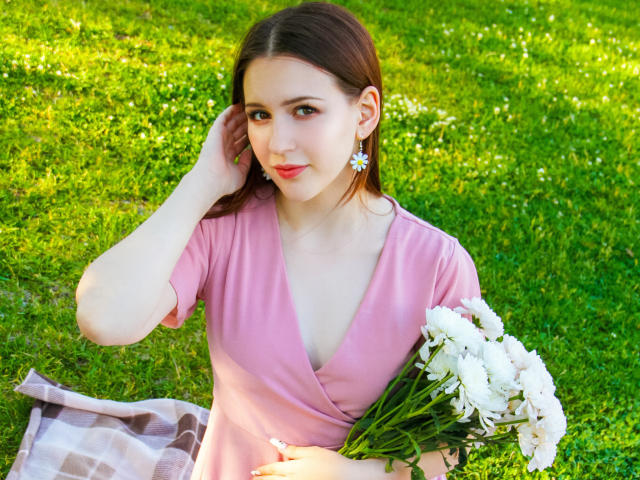 JuliaKing with brown hair, pink dress, and daisy earrings, holding white flowers, sitting on grass, hand in hair, soft smile, fair skin, and cute face, sunny day.