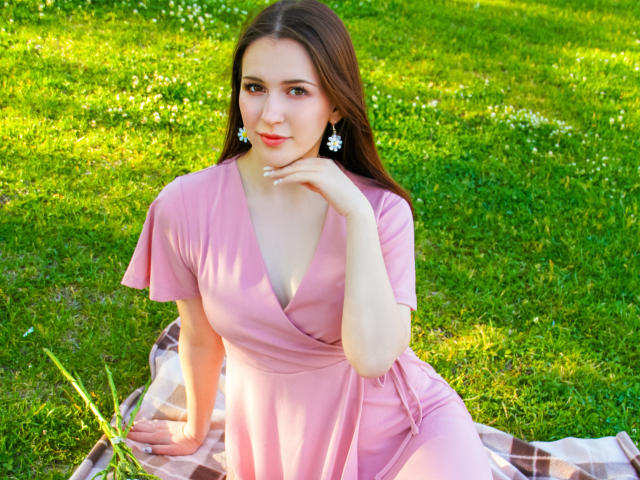JuliaKing with brown hair, pink dress, and white daisy earrings, sitting on a checkered blanket, hand on chin, grassy background, green, summer, cute, tits visible, smiling.