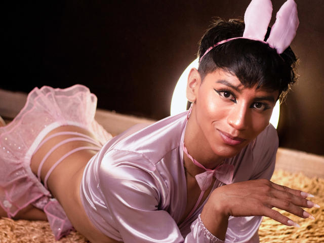 Anddyys with pink bunny ears, lying on straw, wearing a pink satin top, white lace skirt, and pink strappy panties, smiling at the camera. Boobs visible, pussy not shown.