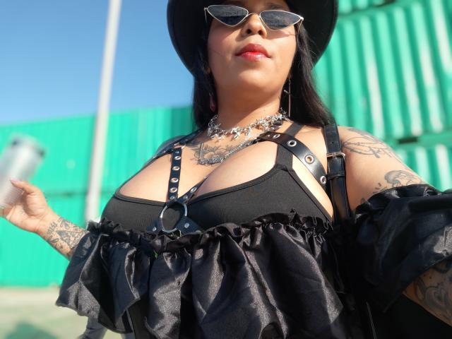 Chili draagt een zwarte off-shoulder top met zilveren studs, zwarte hoed, zonnebril en zilveren ketting, met tatoeages op de armen.