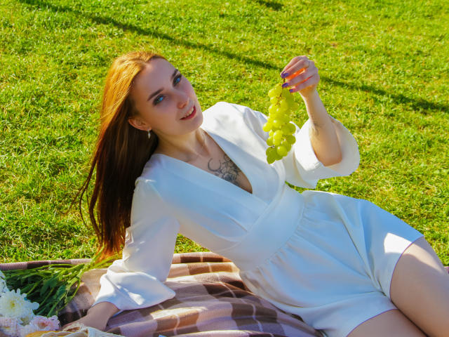AlexisHott with red hair, wearing a white dress, holding green grapes, sitting on a checkered blanket, grassy background, sunny, tattoo on chest, smiling, blue eyes, pink nails.