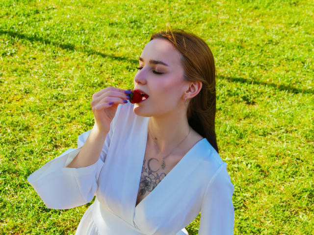 AlexisHott with red lipstick, eating a blueberry, white dress, tattoo on chest, brown hair, green grass, sunlight, holding berry near mouth, cute, sexy.