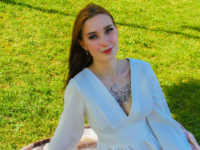 AlexisHott with pale skin, brown hair, white dress, deep V-neck, showing tits, tattoo on chest, sitting on grass, smiling, sunlight, green lawn, red lips, earrings.