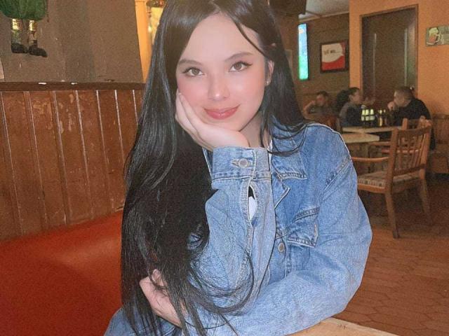 ElyMoon with long black hair, wearing a blue denim jacket, resting her cheek on her hand, smiling softly, sitting in a wooden booth. Her tits are slightly visible.
