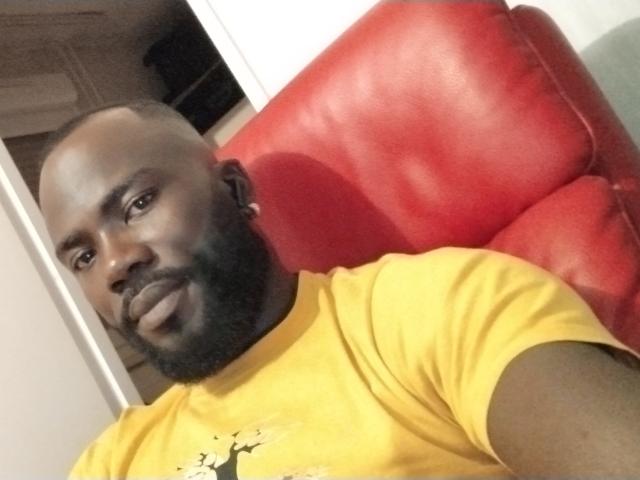 IgorBig with a bald head and black beard, wearing a yellow shirt, sitting on a red leather chair, smiling at the camera. His face is dark-skinned and he's looking relaxed.