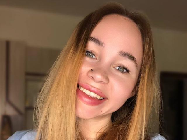 VanillaCake has blonde hair, blue eyes, a small nose piercing, and is smiling with pink lipstick. She's wearing a blue shirt, and her tits are not visible.