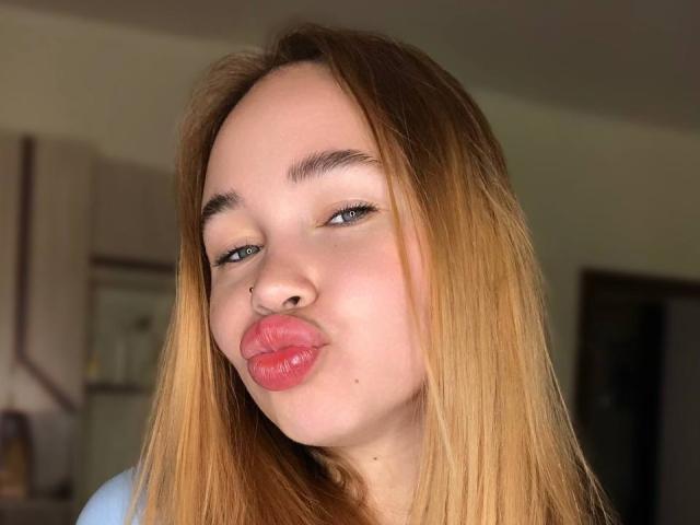 VanillaCake has light brown, straight hair, a nose piercing, and is puckering her pink lips in a kissy face. She has blue eyes and a slight smirk.
