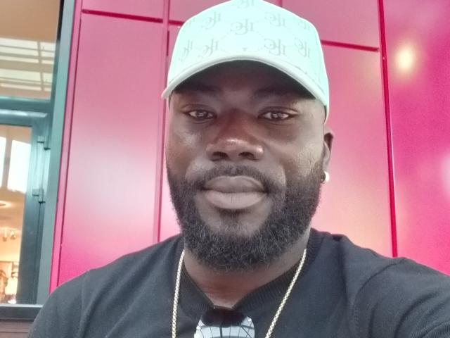 IgorBig, a dark-skinned dude with a full beard, wearing a white cap with text and a black shirt, has a gold chain and a slightly smug face.