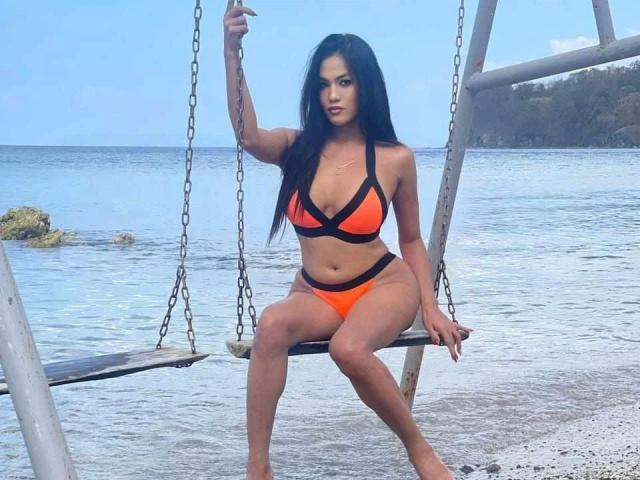 AddictiveMiss4u-hot with long black hair, orange and black bikini, sitting on a wooden swing, holding a chain, blue ocean in background, clear sky, tan skin, curvy figure.