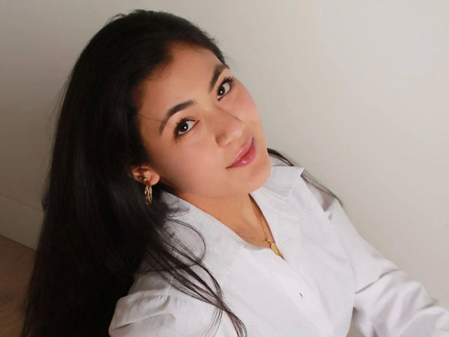 JenniferMet-hot with long black hair, wearing a white shirt, gold earrings, and a gold necklace, smiling softly at the camera, brown eyes, light skin, and pink lips.