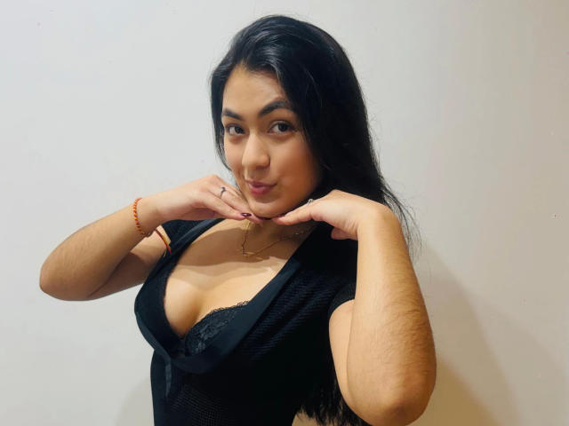 JenniferMet-hot with black hair, medium tits, wearing a low-cut black top, hands near chin, red bracelet, subtle smile, light brown skin, and a necklace.