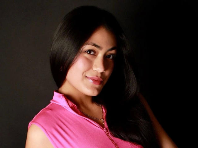 JenniferMet-hot with long black hair, pink zip-up shirt, and a slight smile, showing her cute face and small tits against a dark background.