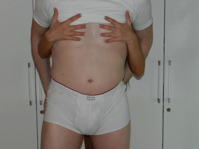 A white-tshirted, white-boxer-wearing hot with hands covering chest, standing in front of a white cabinet. Hot's hands are on their own chest. Hot's penis is not visible.