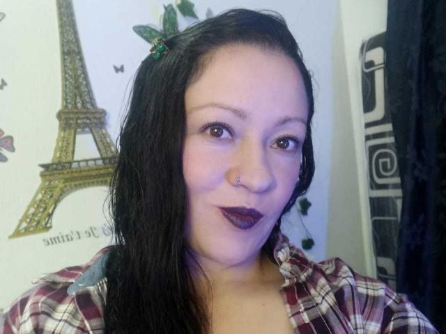 CharloteEzra with wet black hair, nose ring, dark purple lipstick, plaid shirt, and a slight smile, Eiffel Tower wall art behind her.