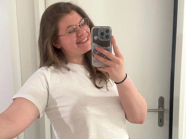 LinaLove - atraente, usando óculos, sorrindo, vestindo uma camiseta branca, tirando uma selfie em um espelho, mostrando seus seios, segurando um iPhone com um suporte pop, em um quarto bem iluminado..