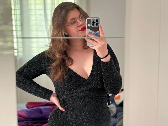 LinaLove - sensual, com batom vermelho, vestindo um vestido preto brilhante, mostrando os seios em uma selfie no espelho, segurando um iPhone, usando óculos, com cabelo castanho ondulado, com a mão na cintura..