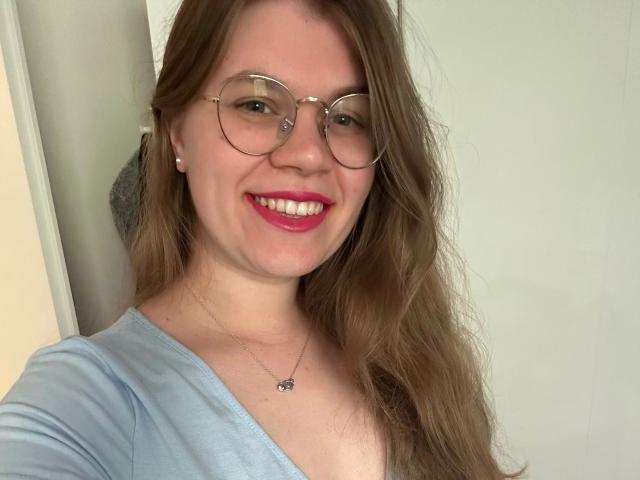LinaLove - uma mulher atraente com cabelos castanhos longos, óculos redondos, batom rosa, sorrindo, camisa azul clara, um colar pequeno e fundo branco. Seios levemente visíveis, nenhuma outra parte do corpo mostrada..