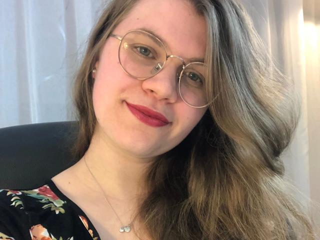 LinaLove, con gafas redondas, lápiz labial rojo y cabello castaño ondulado, sonriendo a la cámara, vistiendo una blusa negra con estampado floral, un collar con una pequeña mariposa y unos jeans muy ajustados..