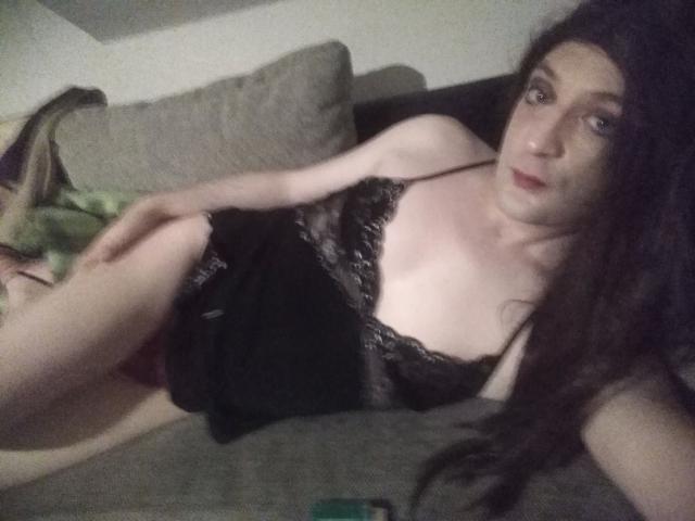 ZackaSissy20-hot, with long brown hair, lying on a grey couch, wearing a black lace camisole, exposing small tits, and showing a pale, slender, sexy body.