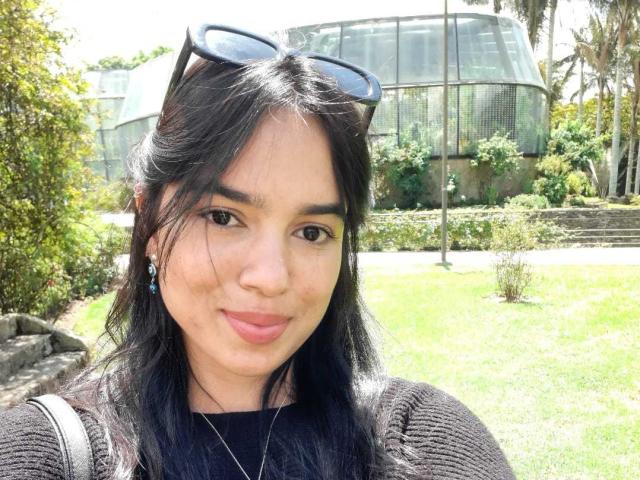 AnyaCathy mit schwarzen Haaren, blauen Ohrringen, rosafarbenen Lippen, einem schwarzen Pullover, einer Sonnenbrille auf dem Kopf, einem grasbewachsenen Hintergrund und einem grünen Glasgebäude im Hintergrund. Ihr Hintern ist nicht sichtbar..