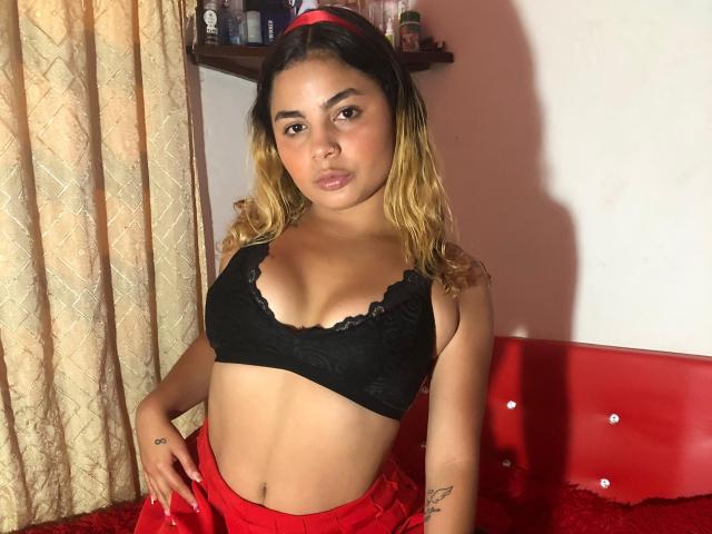JesicaMonics with light brown skin, medium-length wavy blonde hair, red headband, black lace bra, red skirt, tattoos on right arm, standing in front of red bed.