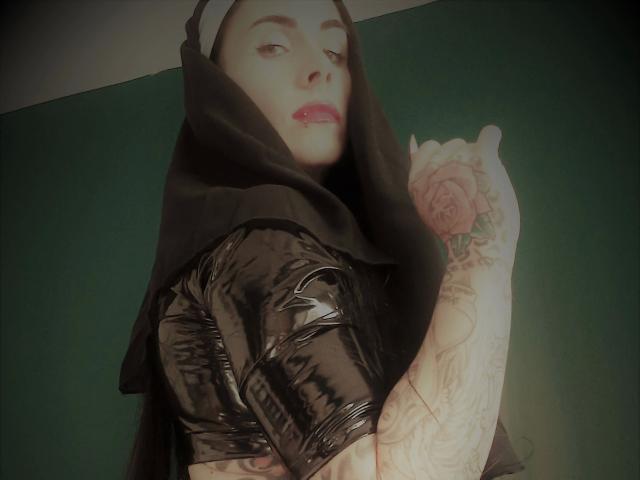 LadyPina-hot with tattoos on her arm, wearing a black nun's habit and shiny black latex top, showing off her rose tattoo, and giving a thumbs-up.