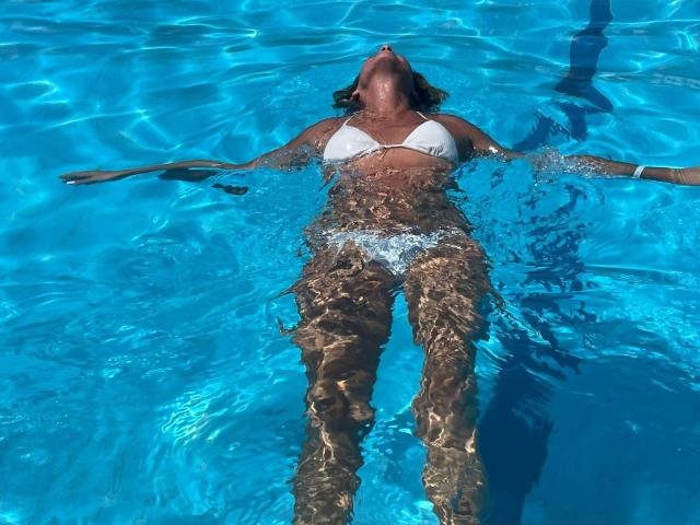 BlackAngeel floating in a blue pool, wearing a white bikini, arms out, head tilted back, water rippling around her legs, sun shining on her tan skin.
