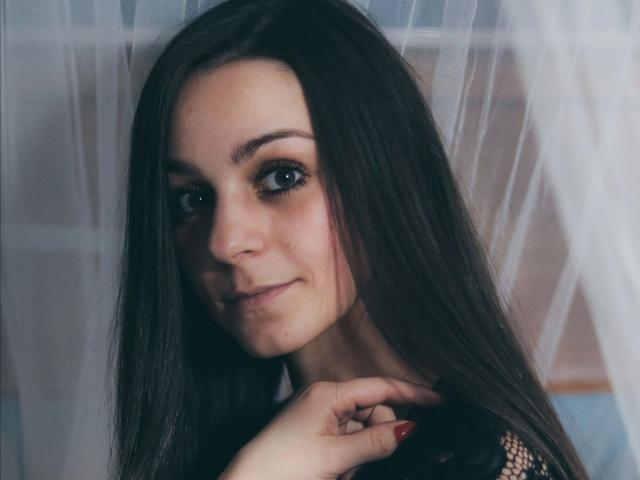 LoellaRose-hot with long brown hair, green eyes, and a slight smile, wearing black fishnet, her hand with red nails touching her neck, soft lighting, subtle makeup.