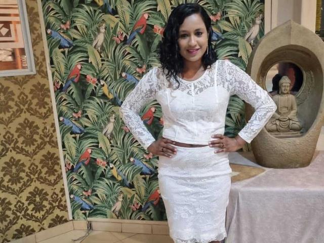 IndainBabelicious, a curvy woman with black curly hair, wears a lace white dress, hands on hips, smiling, standing in front of a colorful tropical-patterned wallpaper.