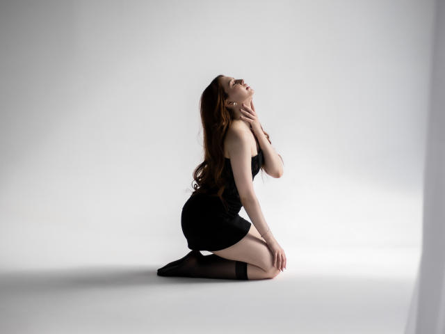 EllyCat, with long brown hair, kneels in black lingerie and stockings, her hand touching her neck, eyes closed, looking up. Her ass is prominent, her pussy is not visible.