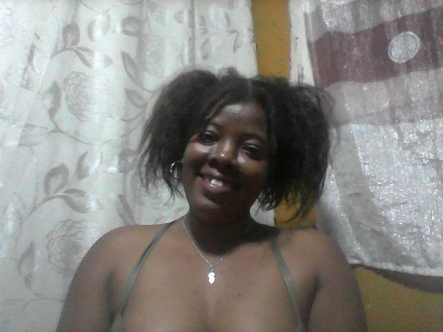BrownieBabe with dark skin and puffy afro, smiling, wearing a green halter top, showing small tits, necklace with heart pendant, in a dimly lit room.