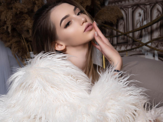 SophieKitty with light brown hair, wearing a fluffy white fur, touching her chin with a hand, looking slightly up with a soft, sexy expression.