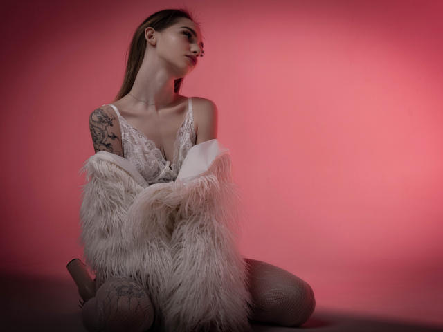 SophieKitty kneeling, wearing lace lingerie, off-shoulder white fur coat, fishnet stockings, tattoos on right arm, pink gradient background, looking to the side, long brown hair.