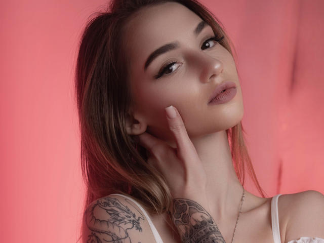 SophieKitty has brown hair, full lips, and tattoos on her right arm. She's touching her face with her left hand, wearing a white top, and has a pink background.