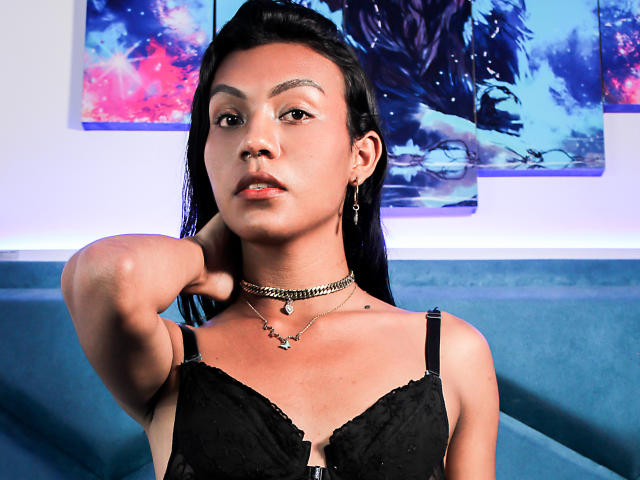KattaleyaHorny with dark hair, black lace bra, and choker, arm raised, looking at camera, wearing earrings, blue couch in background, paintings with blue and pink colors.