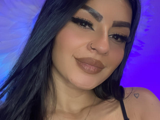 LunaCollinss has black hair, thick eyebrows, long eyelashes, brown lipstick, a nose ring, and a heart tattoo on her right shoulder, smiling with a cute face.